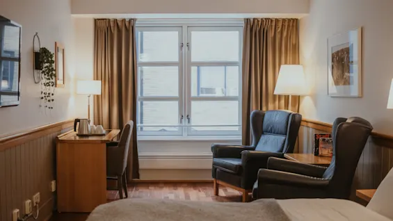 A comfortable hotel room with a bed, a desk, two armchairs, and a large window, creating a cozy and inviting atmosphere.