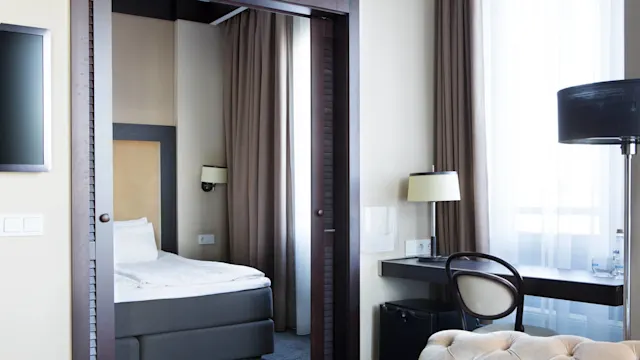 A neatly made bed is visible in a modern bedroom with brown curtains and a desk by the window. A floor lamp and an upholstered chair enhance the cozy atmosphere. Text on a water bottle: "Evian."
