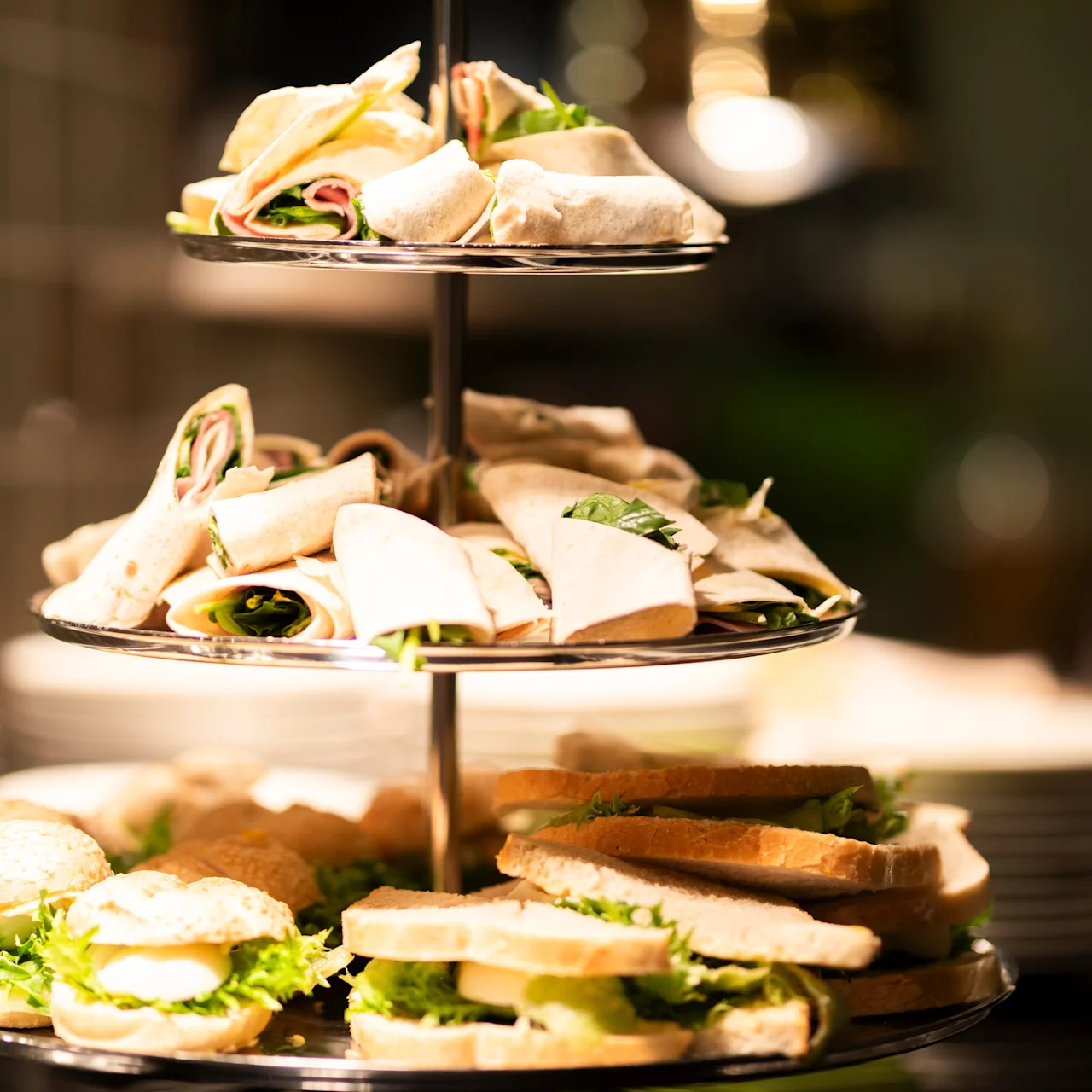 Sandwiches and wraps are stacked on a three-tiered platter. The displays include a variety of fillings, surrounded by a dimly lit buffet setup.