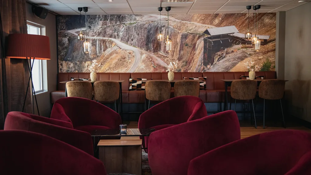 Et komfortabelt salongområde med myke røde lenestoler og benkeseter, med et stort veggmaleri av et ulendt landskap.