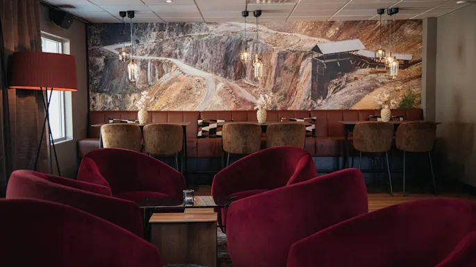 Et komfortabelt salongområde med myke røde lenestoler og benkeseter, med et stort veggmaleri av et ulendt landskap.