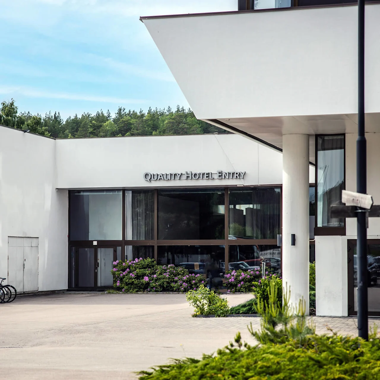 A modern hotel entrance with large glass doors, framed by white walls, features the signs "QUALITY HOTEL" and "QUALITY HOTEL ENTRY". Bicycles and greenery enhance the setting.
