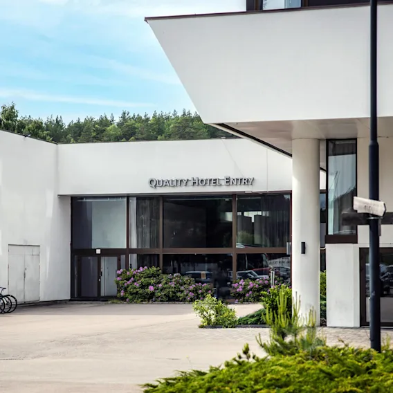 En moderne hotellinngang med store glassdører, omgitt av hvite vegger, har skiltene "QUALITY HOTEL" og "QUALITY HOTEL ENTRY". Sykler og grøntområder forbedrer settingen.