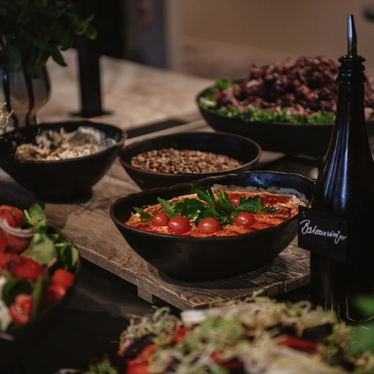 Bowl with cherry tomatoes and greens, arranged on a wooden board surrounded by various salads and dark bottles labeled "Balsamico" and "Olive Oil." Rustic kitchen setting.