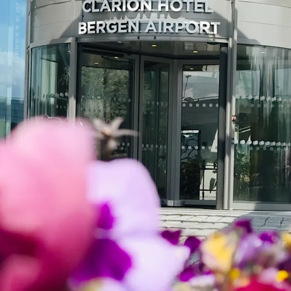 En roterende glassdør merket "Clarion Hotel Bergen Airport" med levende uskarpe blomster i forgrunnen, satt mot en moderne bygning.