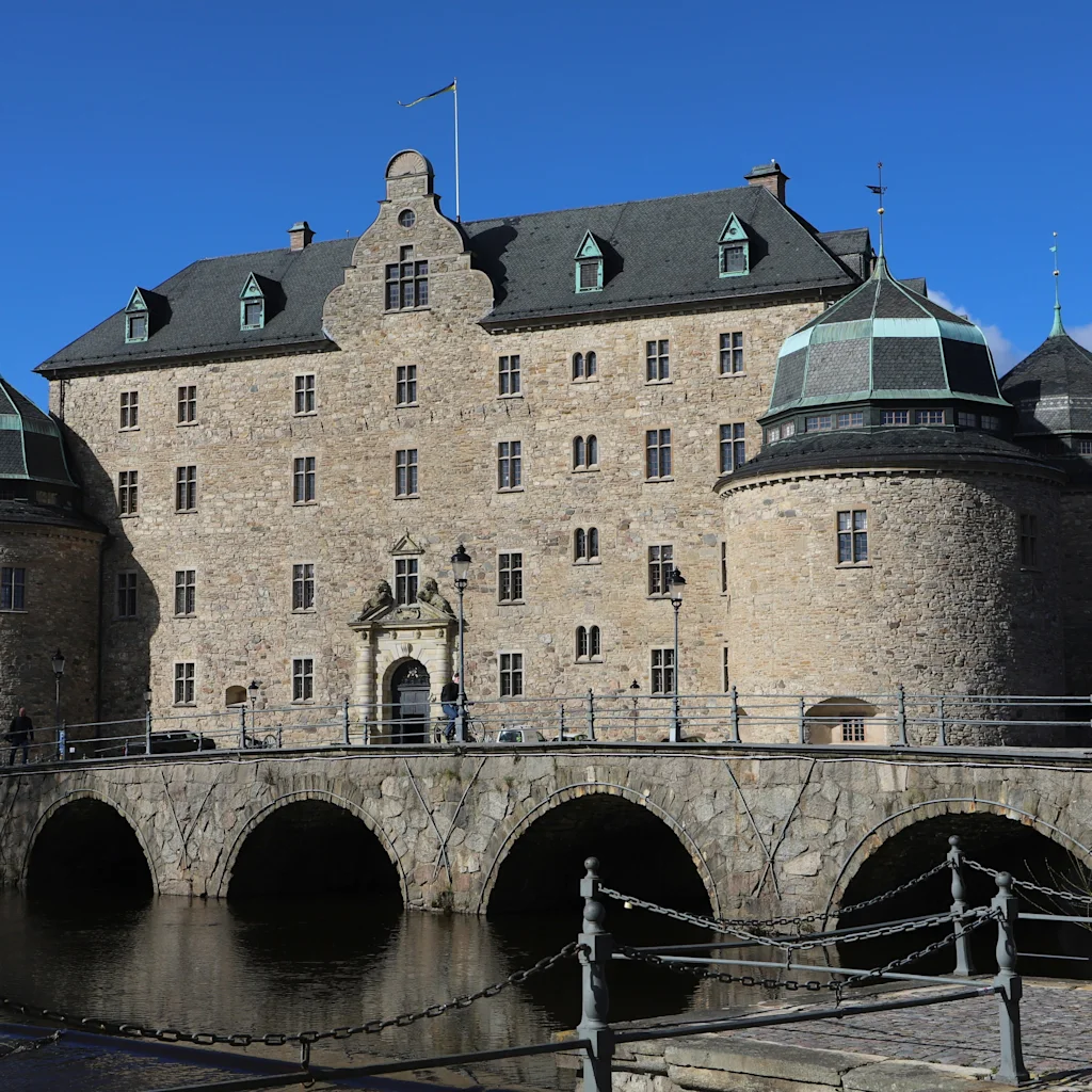 Örebro slott, a grand stone castle with multiple towers, stands majestically over a bridge and water under a clear blue sky.