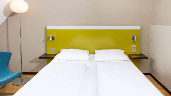 A neatly-made bed with white linens rests against a vibrant green headboard in a minimalist room; a sculptural lamp stands beside a blue chair on wooden flooring.