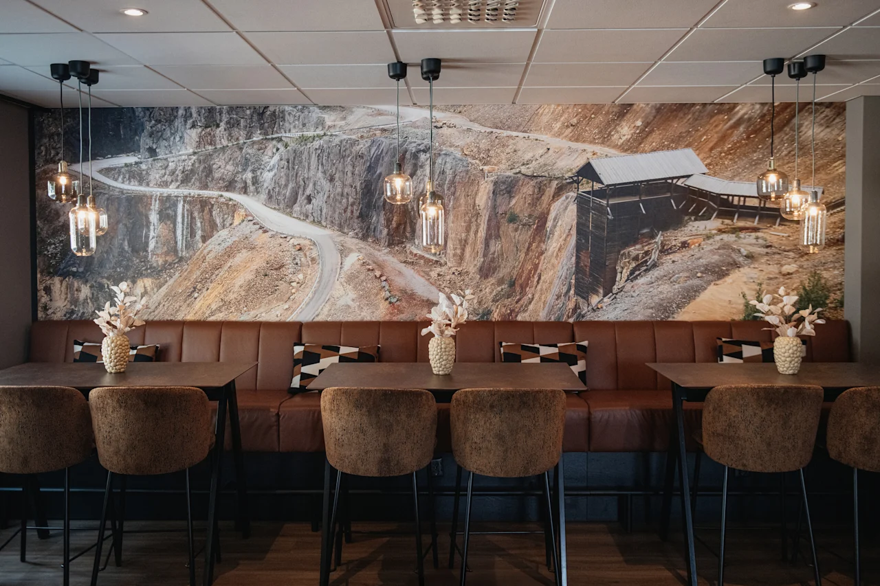 Et moderne spiseområde med brune skinnbenker, høye bord og stoler, med et stort veggmaleri av en svingete fjellvei.