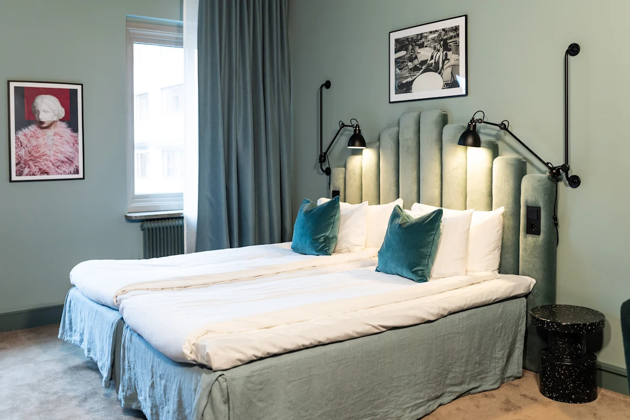 Viihtyisä hotellihuone kahdella sängyllä, pehmeällä valaistuksella ja rauhoittavilla vihreillä ja sinisillä sävyillä, kutsuen rentoutumaan.