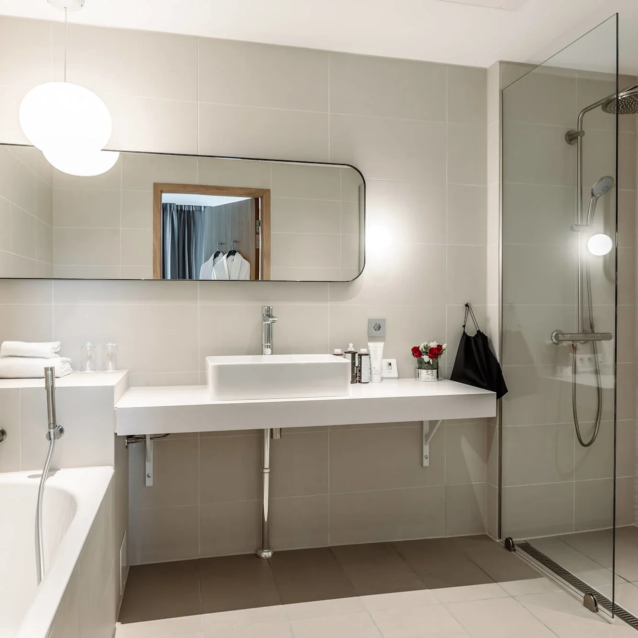 A modern bathroom features a bathtub with a showerhead, a spacious countertop with a sink, towels, and toiletries. A glass-enclosed shower stands adjacent, all set in a sleek, tiled room.
