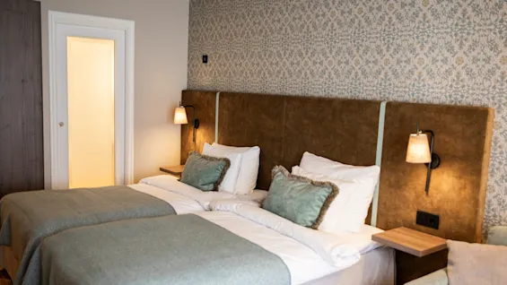 Två prydligt bäddade enkelsängar med ljusblå överkast och gröna kuddar är placerade i ett hotellrum med mönstrad tapet och individuella sänglampor på de vadderade sänggavlarna.