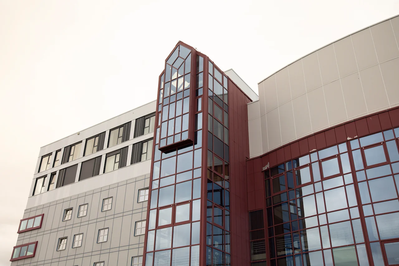 Moderni arkkitehtoninen rakennus, jossa on suuri, monikerroksinen lasiosa, joka ulottuu ulospäin, täydennettynä punaisilla ja harmailla paneeleilla.