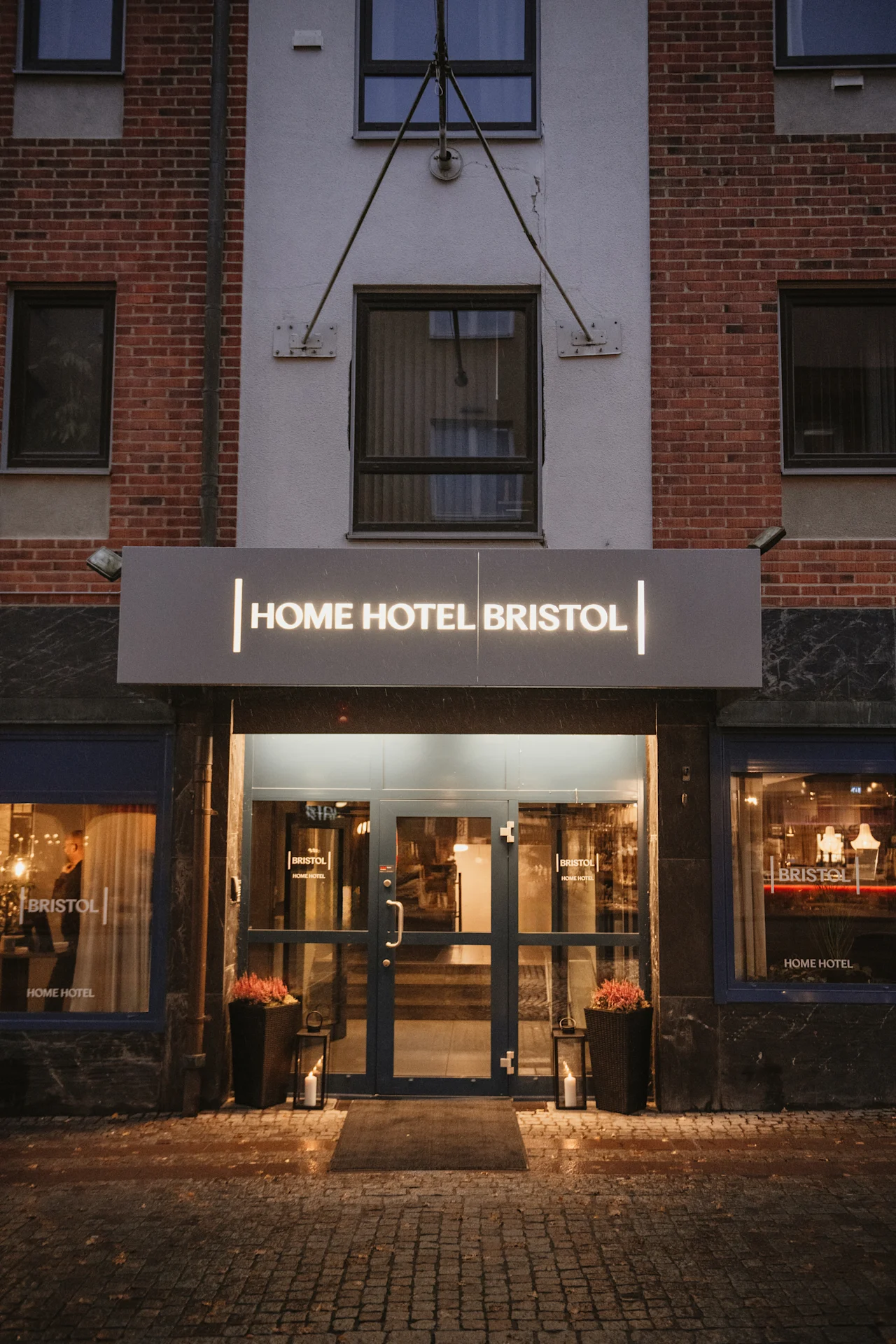 Hotellin valaistu sisäänkäynti illalla, jossa kyltti HOME HOTEL lasiovien yläpuolella ja märkä mukulakivikatu.