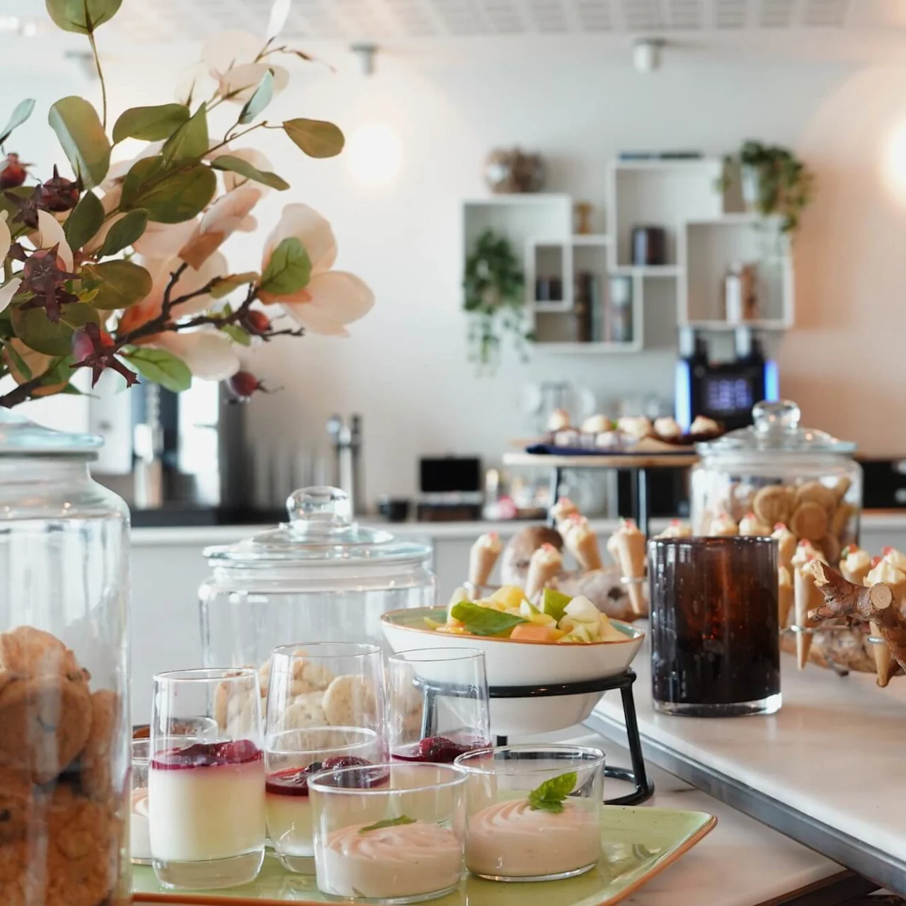 Ornamental arrangement of assorted desserts on a table; colorful pastries, cookies in jars, and decorative foliage present in a bright, modern dining space.