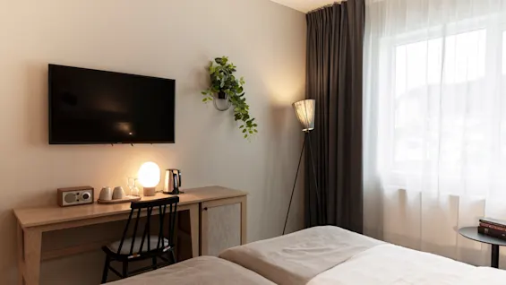 Viihtyisä hotellihuone, jossa kaksi sänkyä, työpöytä TV:llä ja ikkuna ohuilla ja tummilla verhoilla, luoden rentouttavan tunnelman.