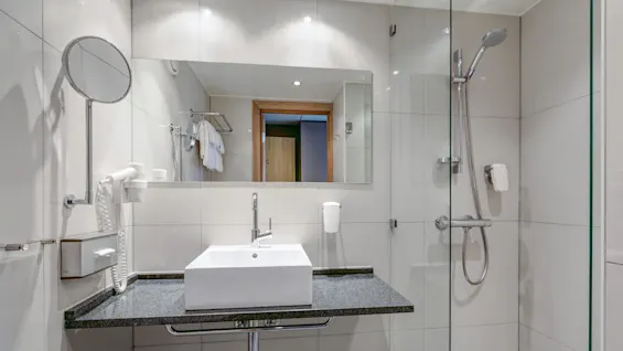 A modern bathroom features a rectangular sink and faucet on a granite counter, with a circular mirror and shower area; towels hang on a rack and white tiled walls surround.