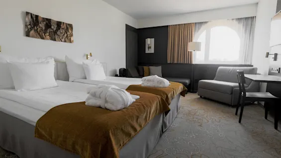 Two neatly made twin beds with white linens and brown throws stand in a modern, well-lit hotel room. The room includes a sofa, desk, and large window with curtains.