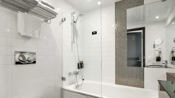 A modern bathroom features a white tiled bathtub with a showerhead, adjacent toilet, and mirrored wall, creating a clean and bright environment.