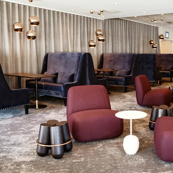 Chairs are arranged around small tables in a stylish lounge with plush seating, dark curtains, and modern spherical pendant lights.