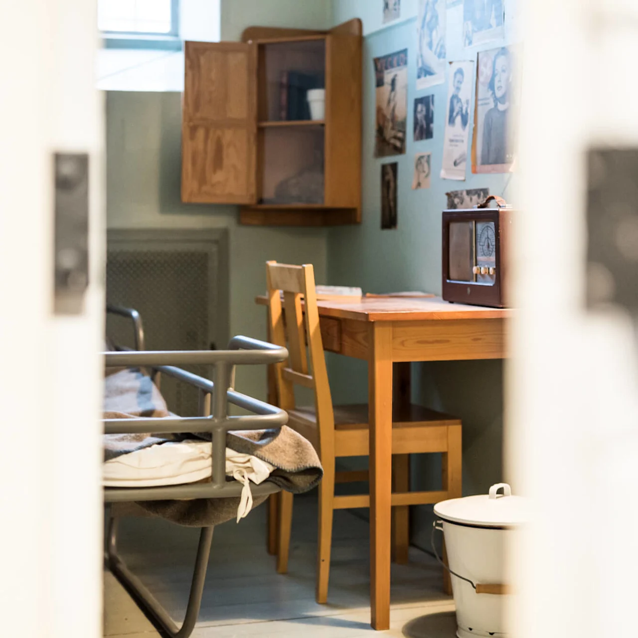 View of a vintage room through an open door, featuring a wooden table and chair, a bed, and a small TV set, with pictures on the wall.