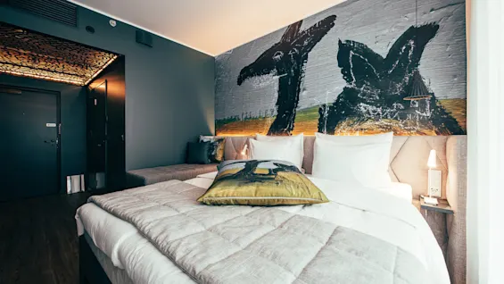 A modern hotel room with a bed featuring white linens and a large mural of abstract black animal figures on a gray wall, accompanied by warm lighting and minimalist furniture.