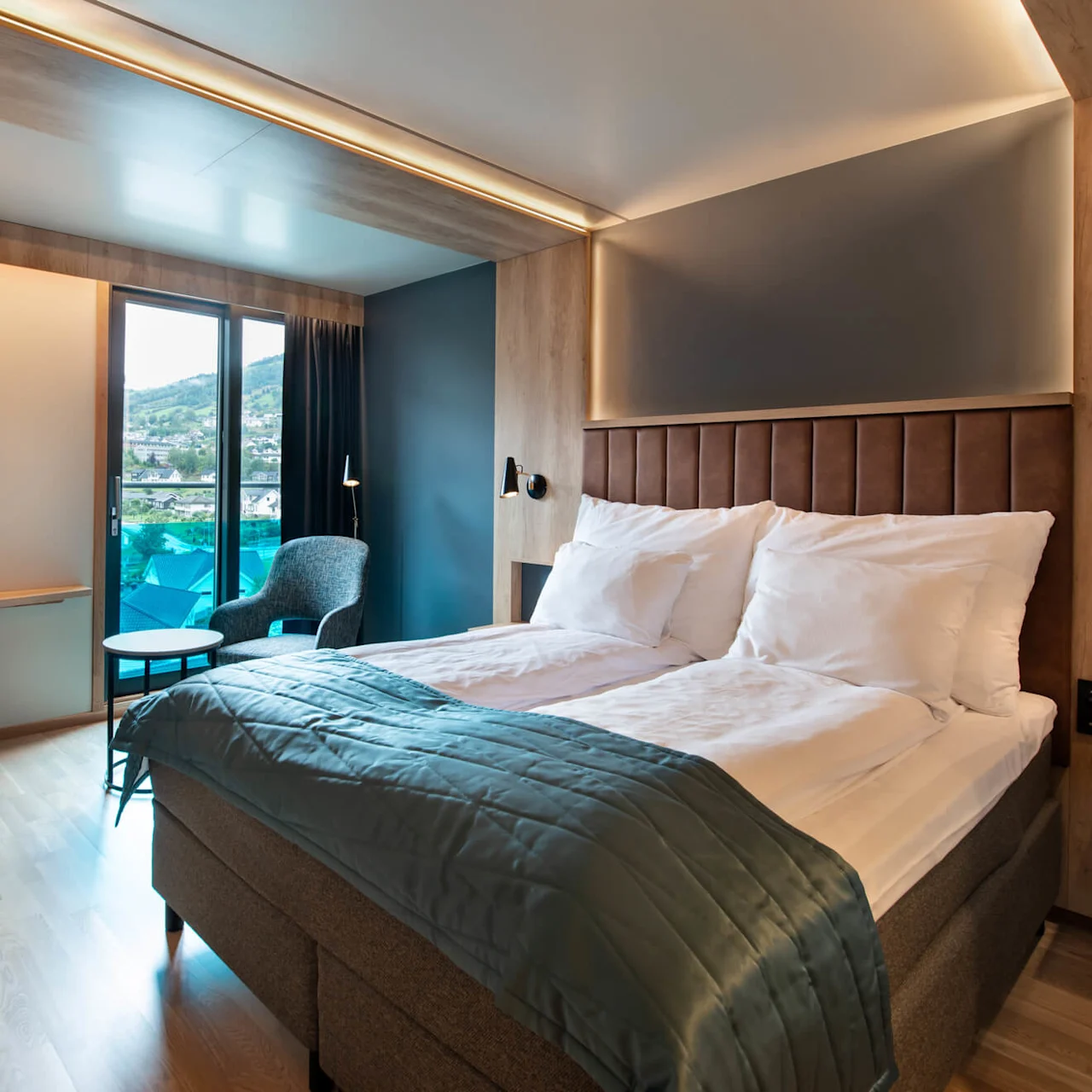 En prydligt bäddad säng med vita kuddar och ett blått täcke står i ett modernt hotellrum. Rummet har ett skrivbord, stol, rund spegel och utsikt genom glasdörrar.