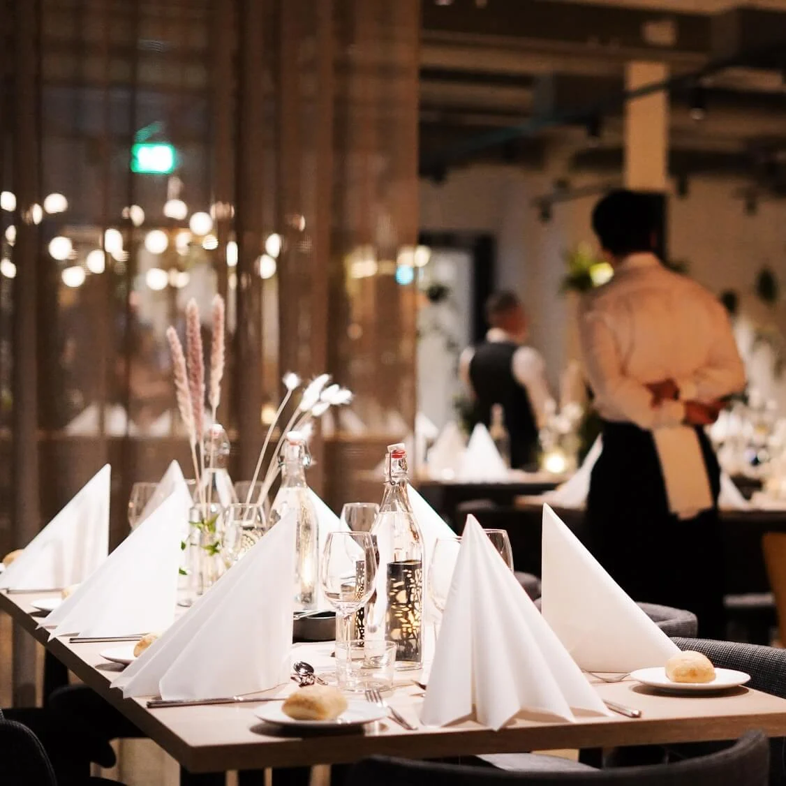 A set dining table with folded napkins and bottles, set in a dimly lit restaurant. A server stands in the background, hands clasped, adding to the elegant ambiance.