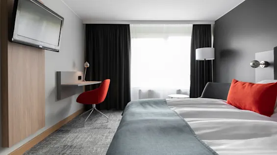 En säng med blått överkast och orange kuddar står bredvid ett modernt skrivbord med en röd stol, placerat mot ett fönster med mörka gardiner i ett hotellrum.
