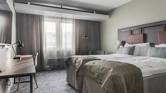 Moderni hotellihuone, jossa kaksi sänkyä, työpöytä ja ikkuna verhoilla, tarjoten mukavan majoituksen.