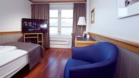 Et ryddeligt hotelværelse med en seng, tv der viser et program, lænestol, skrivebord, og vindue med udsigt til bjerge.