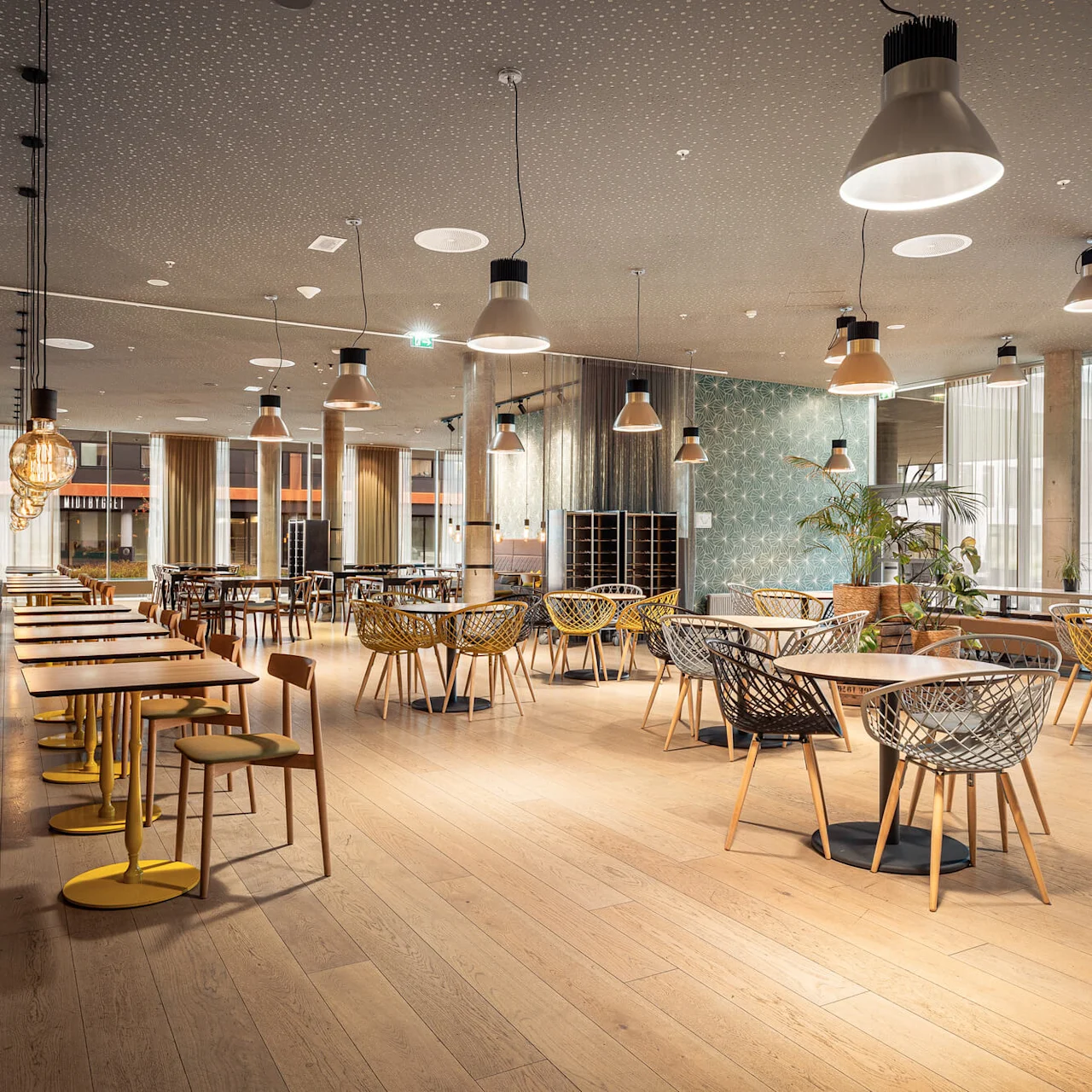 Café interior features modern chairs and tables with hanging lamps, arranged in a spacious, well-lit room with large windows and decorative wall designs.