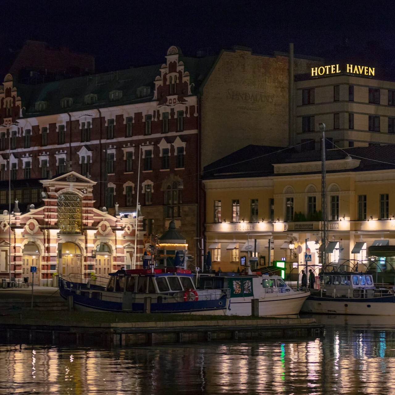 Boats rest docked at night, casting reflections on the water. Illuminated historic buildings are in the background. Signs read "HOTEL HAVEN" and "SUNDSTRÖM."