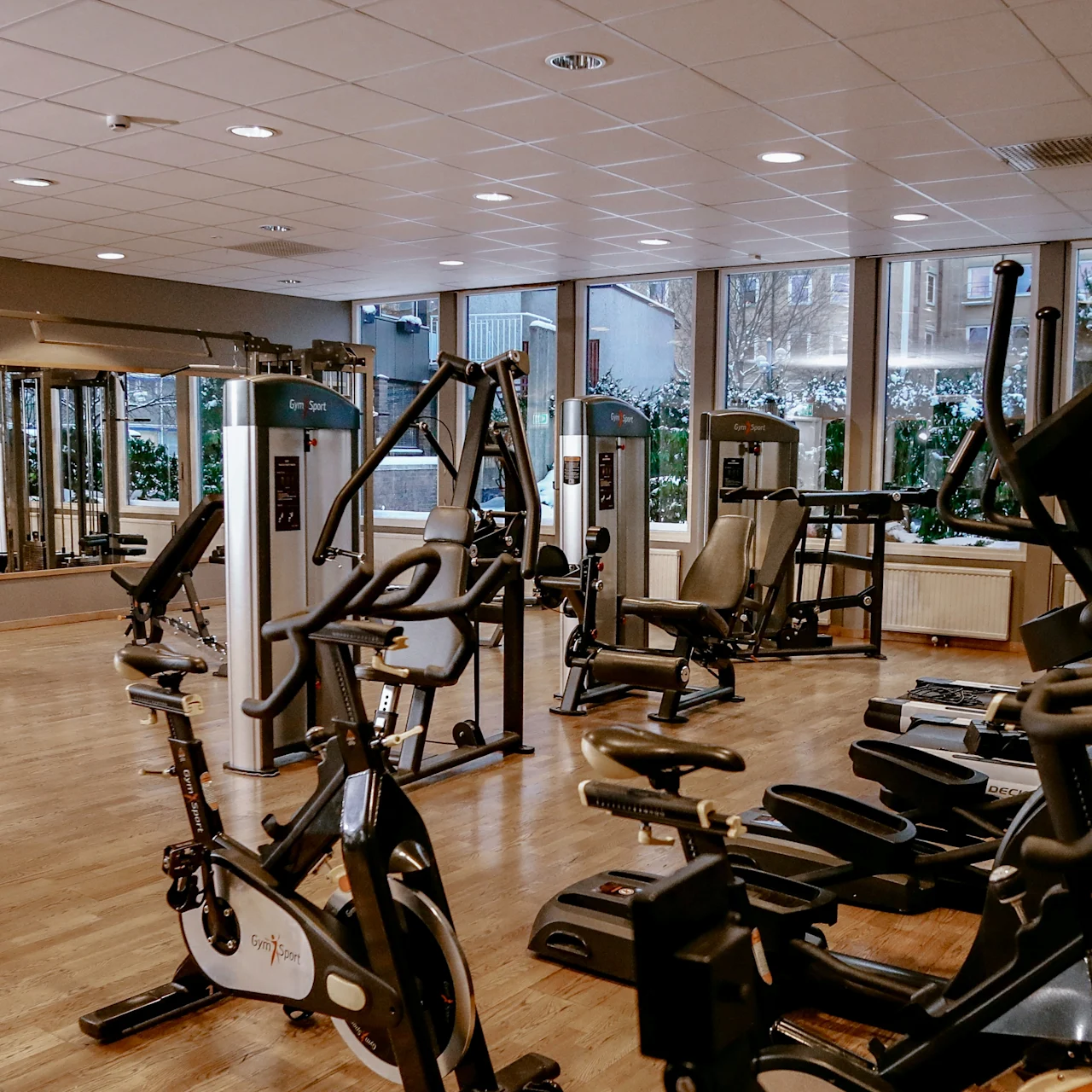 A gym with various exercise machines stands empty. The space is well-lit with large windows, reflecting a clean and organized environment. Text: "Gym|Sport".