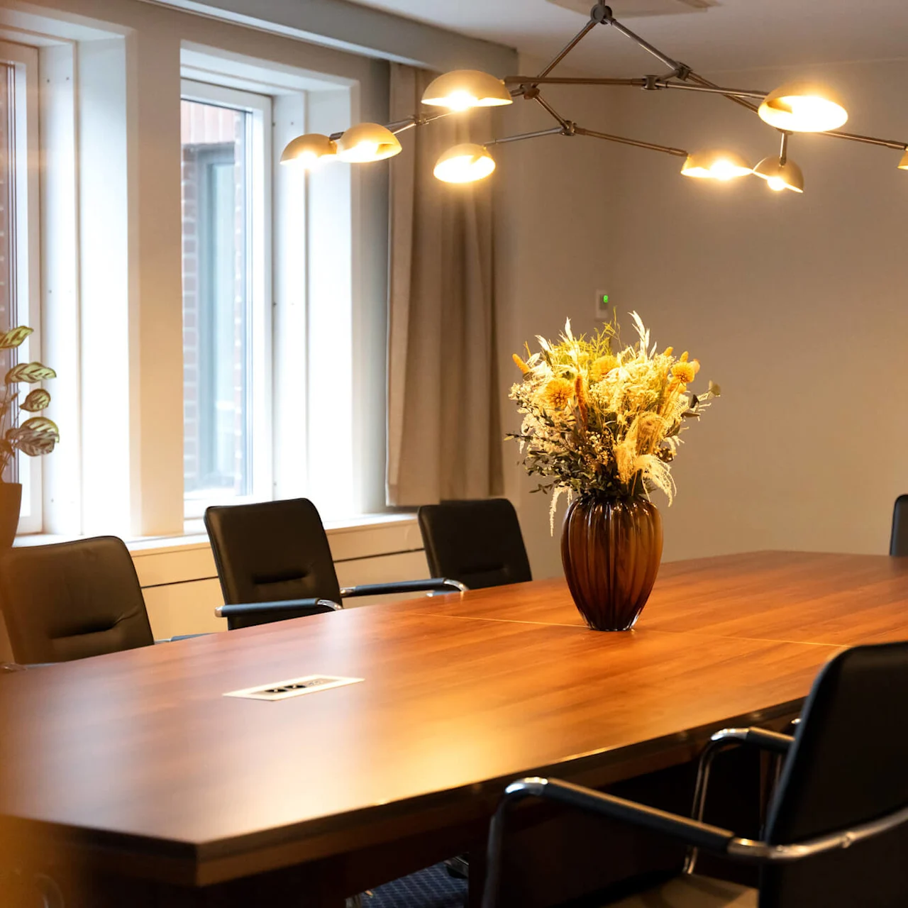 En vas med torkade blommor står på ett konferensbord av trä omgivet av svarta stolar. Taklampor lyser upp rummet, som har stora fönster och en växt i hörnet.