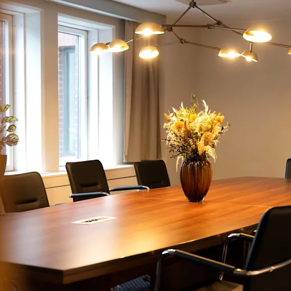 En vas med torkade blommor står på ett konferensbord av trä omgivet av svarta stolar. Taklampor lyser upp rummet, som har stora fönster och en växt i hörnet.