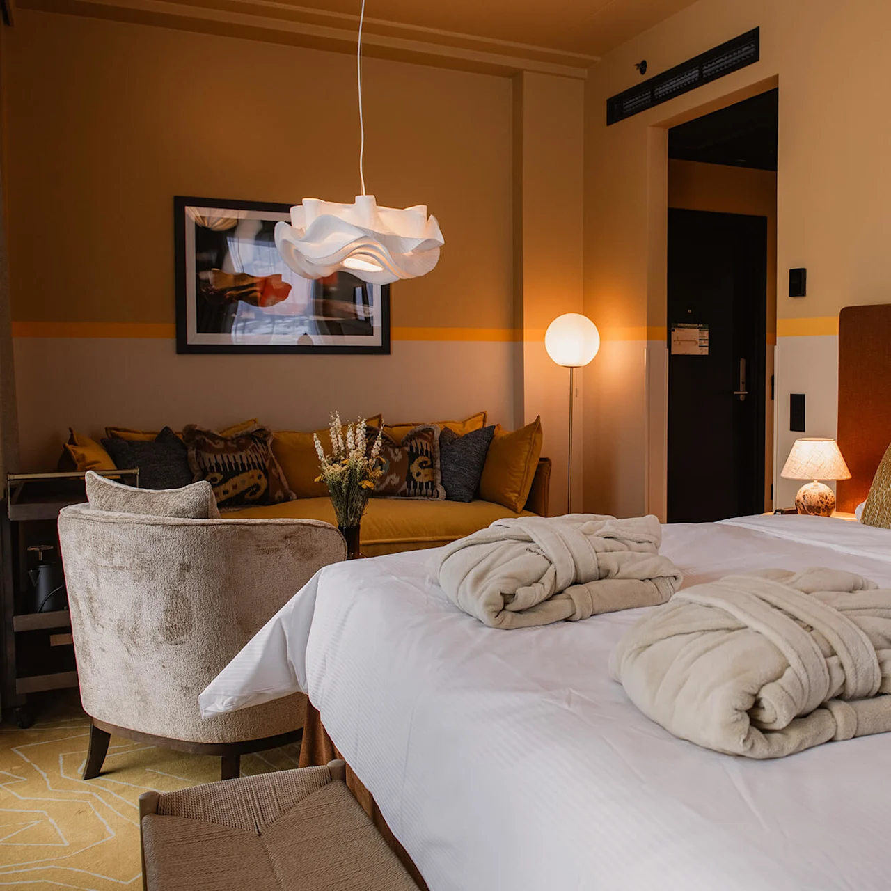 Ett bekvämt hotellrum med en prydligt bäddad säng och två vikta morgonrockar, omgiven av varm belysning och stiliga möbler, skapar en mysig och inbjudande atmosfär.