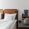 A neatly made bed with white bedding next to a wooden nightstand featuring a phone, a lamp, and an information card in a tidy hotel room.