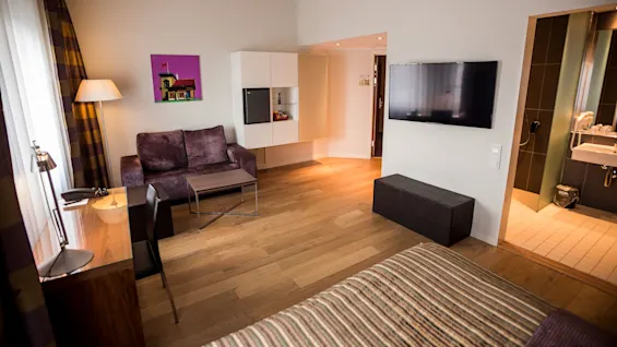 Et moderne hotellrom med sofa, skrivebord og veggmontert TV. Ved siden av er et lite bad. Parkettgulv og myk belysning skaper en hyggelig stemning.