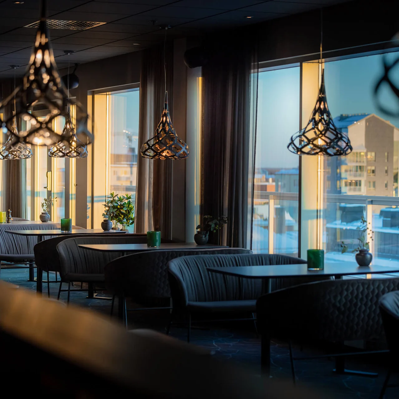 Stiliga hänglampor hänger ovanför bekväma sittplatser i en elegant, modern restauranginteriör, solljus strömmar genom stora fönster med utsikt över stadens byggnader.