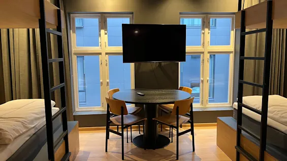 Moderni hotellihuone, jossa kerrossängyt, pyöreä ruokapöytä tuoleineen, seinä-TV ja suuret ikkunat.