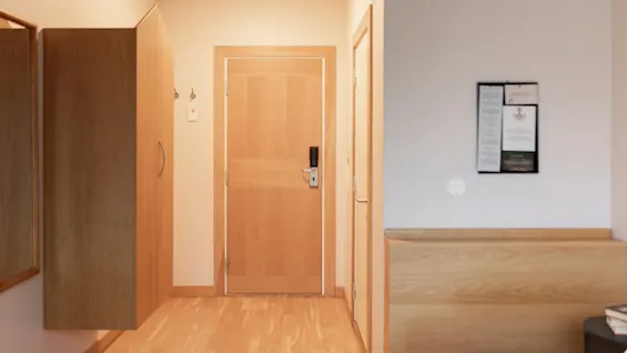 En lys hotellromsinngang med tredør, garderobe og oppslagstavle på veggen, som fører inn til soveområdet.