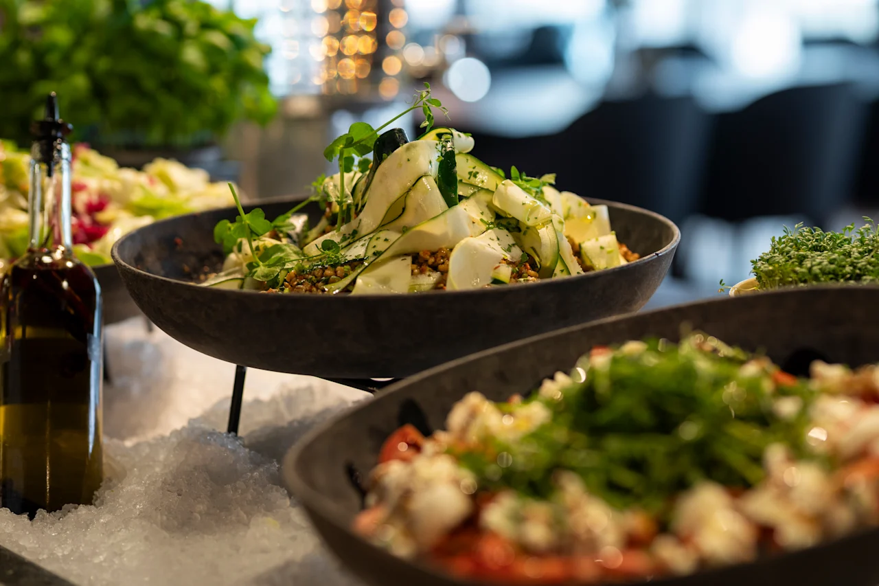 En skål med färsk sallad med skivade grönsaker och örter vilar på en bädd av is, omgiven av andra rätter, inomhus med mjuk belysning.
