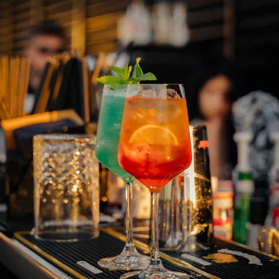 Two vibrant cocktails, one blue and one orange, sit on a bar, ready to be enjoyed in a lively atmosphere.