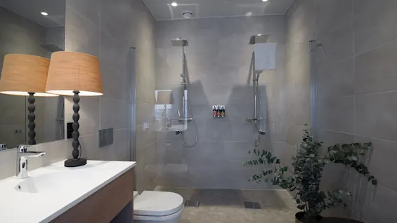 A modern bathroom with beige tiles features a dual shower setup, a toilet, a wooden vanity with a lamp, and a pot with green foliage, creating a contemporary ambiance.