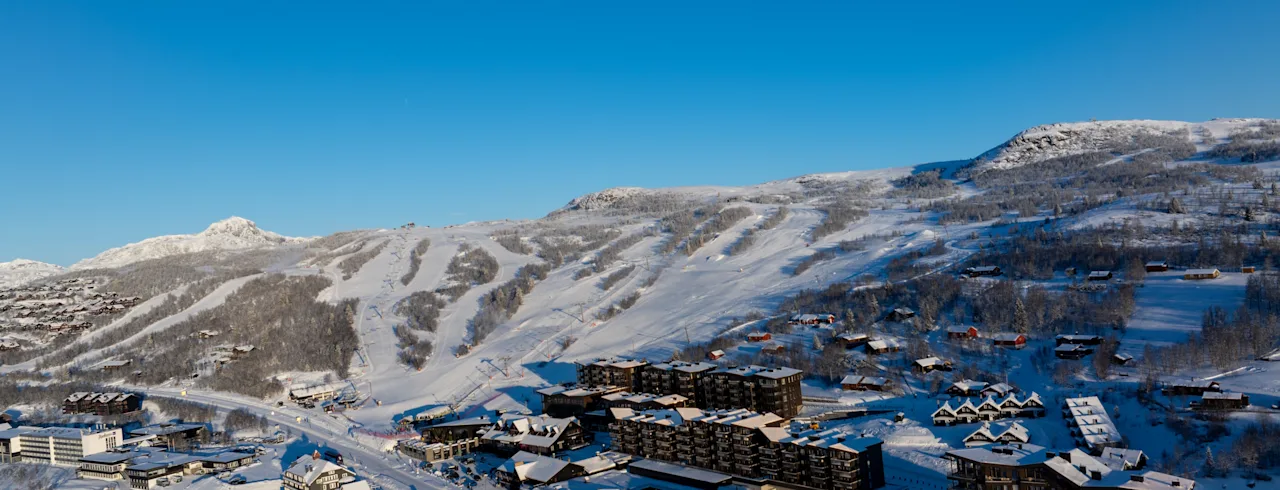 En flygbild över en snötäckt skidort med byggnader inbäddade bland bergen och synliga skidbackar under en klarblå himmel.