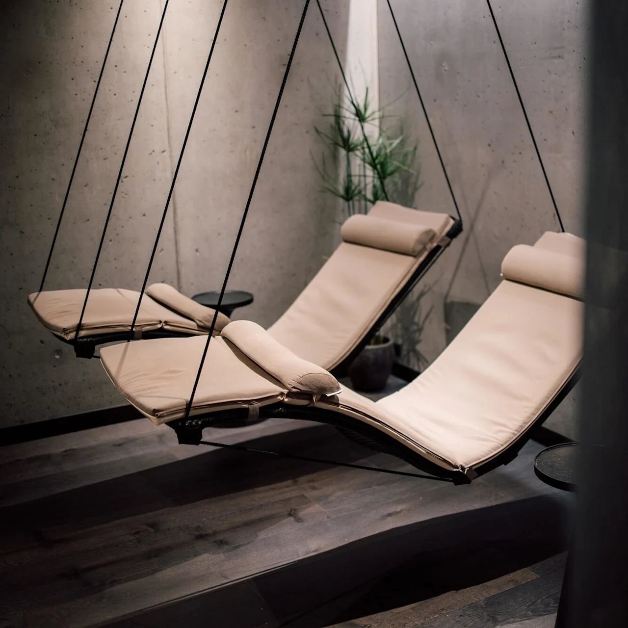 To beige liggestole, ophængt fra loftet, hænger i et minimalistisk rum med mørkt trægulv og betonvægge. Et lille sidebord og en potteplante er i nærheden.