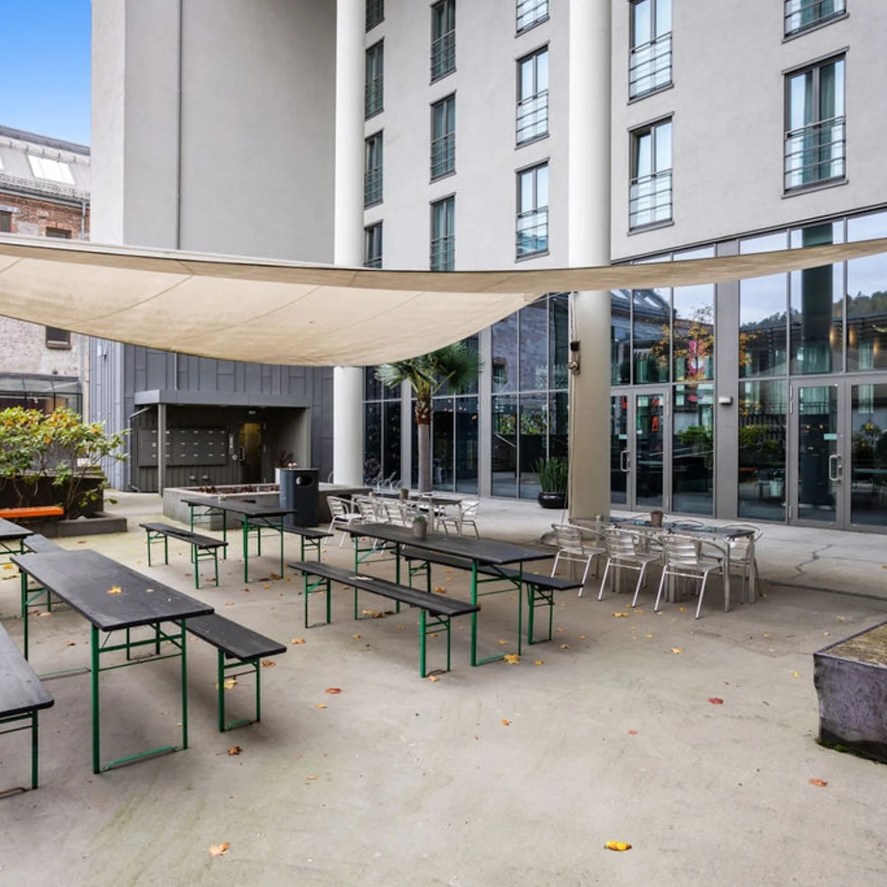 Outdoor courtyard with green-framed tables and benches under a beige sunshade, adjacent to a modern grey building with tall windows and glass doors. Sparse vegetation surrounds the area.