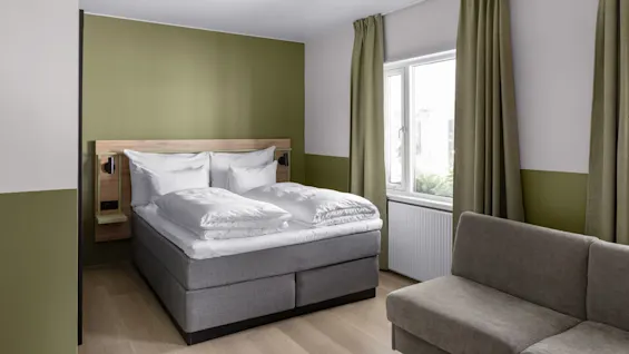 En prydligt bäddad dubbelsäng med vita sängkläder står mot en olivgrön vägg, bredvid ett fönster med gröna gardiner, i ett modernt sovrum med en grå soffa.