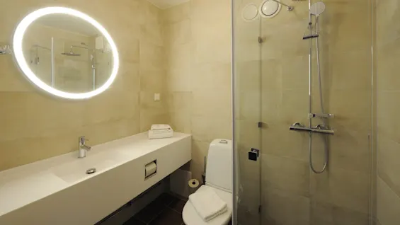 A modern bathroom features a round, illuminated mirror above a white sink, adjacent to a toilet and glass-enclosed shower. Beige tiles cover the walls, creating a minimalist atmosphere.