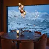 A round dining table, set with glasses and cutlery, under warm hanging lights. Beyond a large window, snow-covered mountains and chalets create a serene winter landscape.